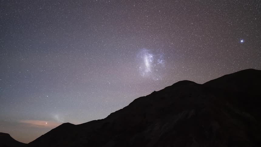 A breathtaking night-to-day timelapse shows the Large and Small Magellanic Clouds setting over a desert mountain as the landscape is illuminated by the rising sun