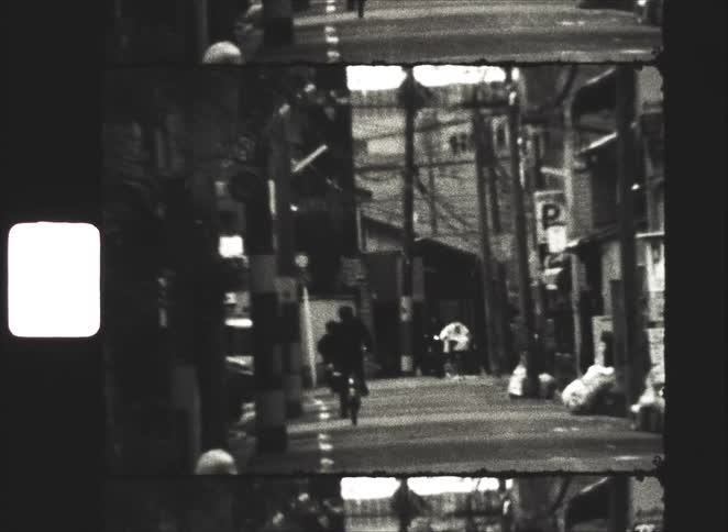 Vintage 8mm black and white footage shows a person riding a bicycle down a narrow urban street in Japan