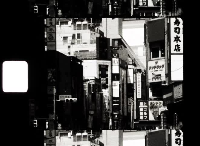 A gritty, black and white vintage film clip slowly zooms in on a narrow, busy street in a Japanese city, filled with signs and buildings