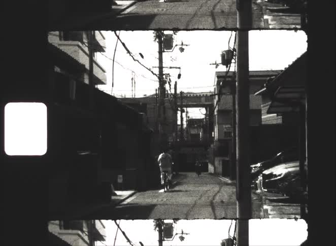 A vintage, black and white 8mm film shows a cyclist and a scooter riding away down a narrow urban alley in Japan