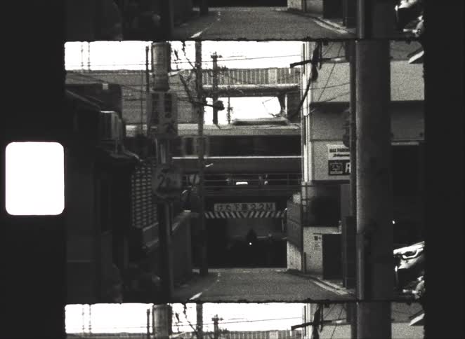 A gritty black and white Super 8mm film captures a train passing over a narrow urban alley in Japan