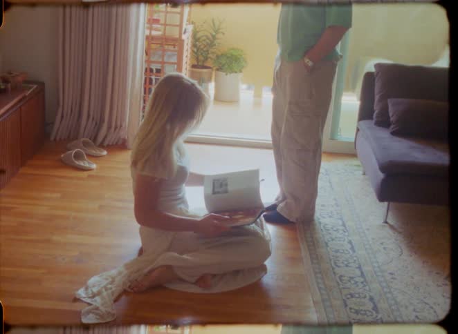 Vintage-style footage shows a woman in a white dress reading on the floor and looking up at a man in a sunlit living room