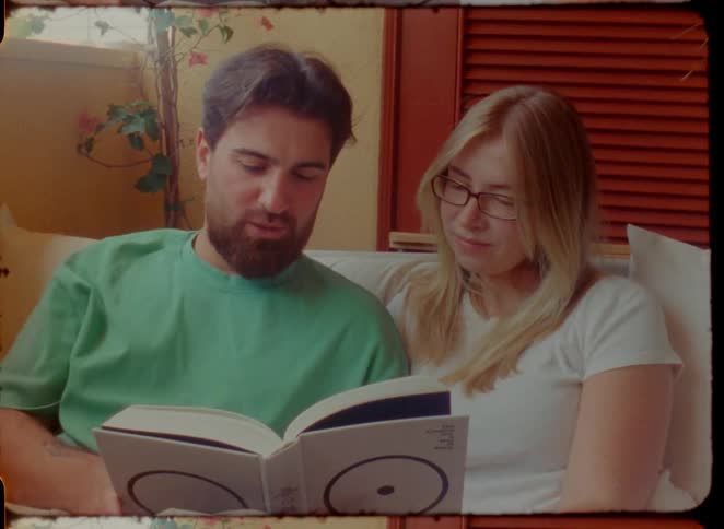 A romantic couple reads a book together on the couch before sharing a sweet kiss in this intimate, vintage-style footage