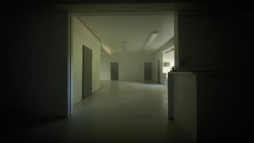 A foreboding static shot from a dark room peers into an empty, sterile hallway of an abandoned institution
