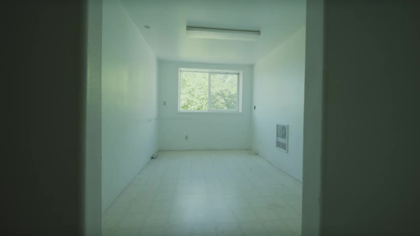 A fixed, unsettling view of a stark, empty white room with a single window revealing trees outside
