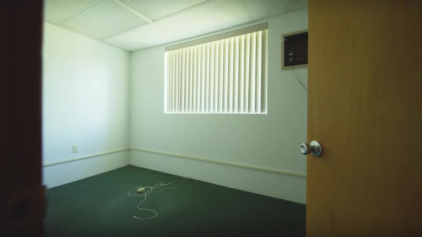 A static shot of an empty, vacant office with white walls, green carpet, and a window with blinds