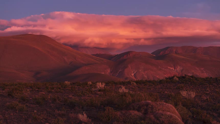 A day-to-night timelapse of dramatic pink clouds rolling over a vast, arid mountain range as the sky transitions from sunset to a starry night