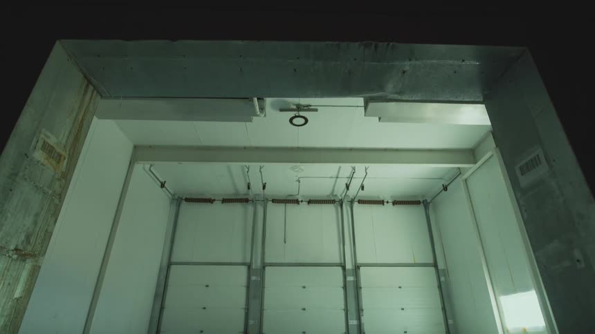 Tilting down from a dark entryway to reveal a large, empty industrial loading bay with three white doors