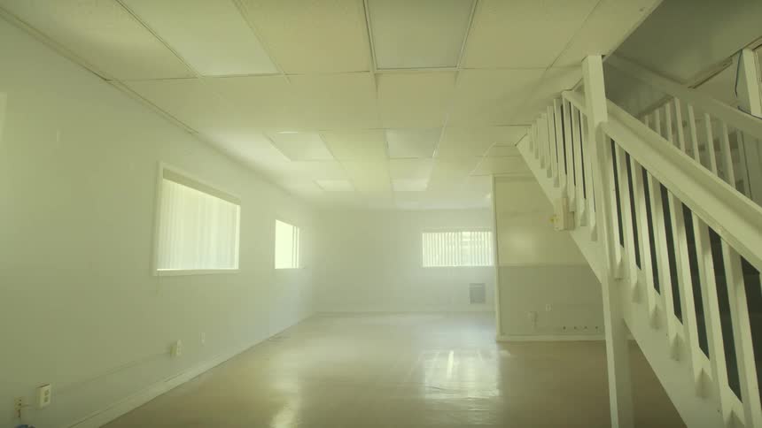 Tilting down from a drop ceiling to reveal a large, empty, unfurnished basement with windows and a white staircase