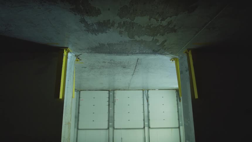 Slowly zooming out from a dark, weathered concrete ceiling to reveal three large white loading bay doors in an empty industrial warehouse