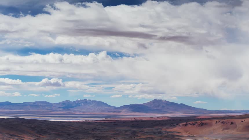 A dramatic timelapse of clouds moving swiftly over a vast, arid mountain landscape, casting shadows across the red desert floor and a distant salt flat