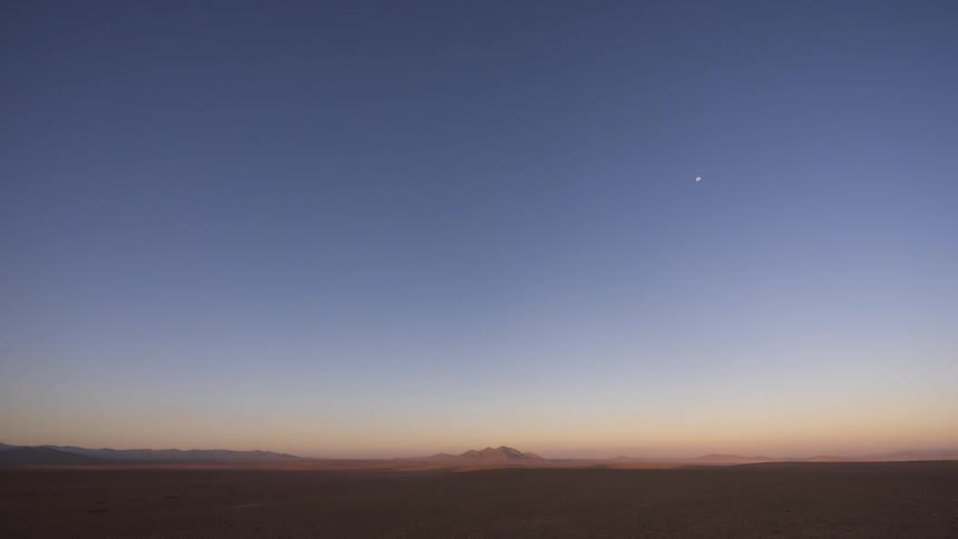 A time-lapse captures the moon setting and the sun rising over a vast and barren desert, transitioning the landscape from deep night to bright day