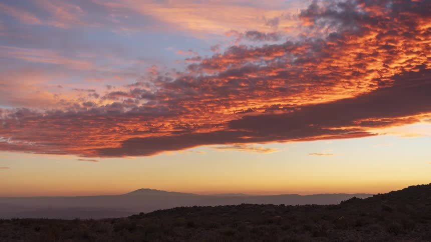 A stunning time-lapse captures a dramatic sunset as clouds blaze with fiery orange and red hues over a distant mountain silhouette