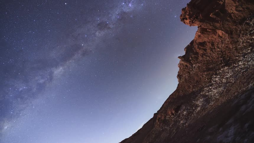 A stunning time-lapse captures the Milky Way galaxy arching across the night sky above a dark, rocky desert landscape, with the camera slowly tilting upwards