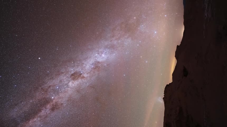 A vertical time-lapse captures the stunning Milky Way galaxy as it moves across the night sky, transitioning to a brilliant sunrise over a rocky landscape