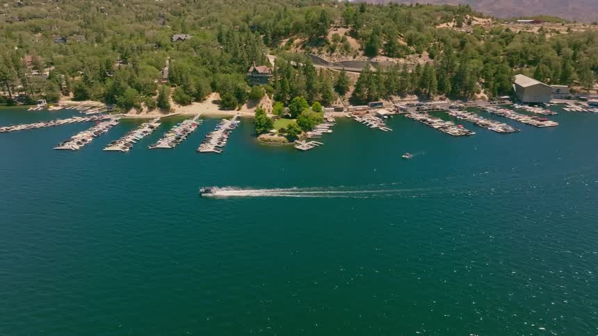 Aerial drone footage follows a speed boat cruising across a beautiful mountain lake past a marina filled with docked boats