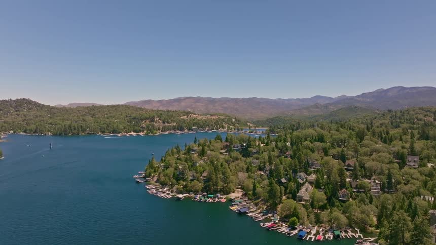 Sweeping aerial footage captures the stunning blue waters of a mountain lake, lined with charming houses and boat docks nestled in a dense green forest