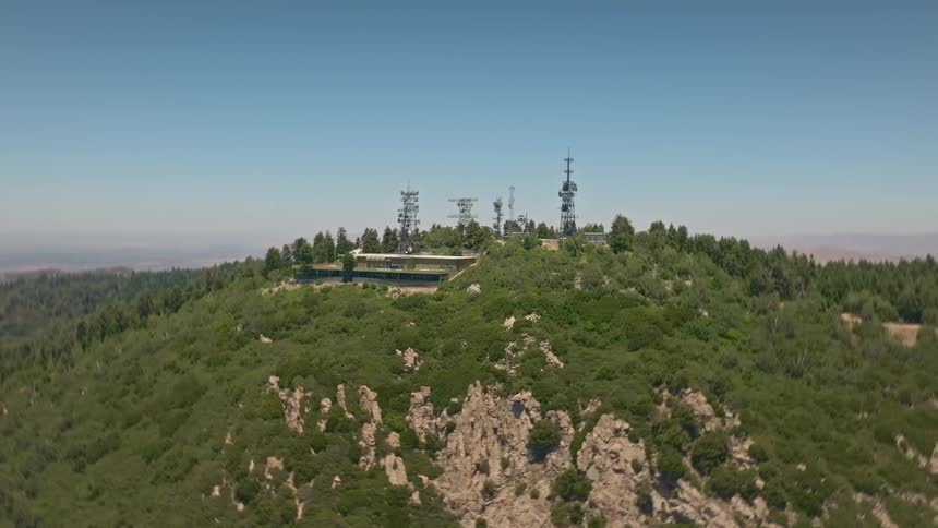 An aerial shot slowly pulls back to reveal a cluster of communication towers on a verdant, rocky mountaintop