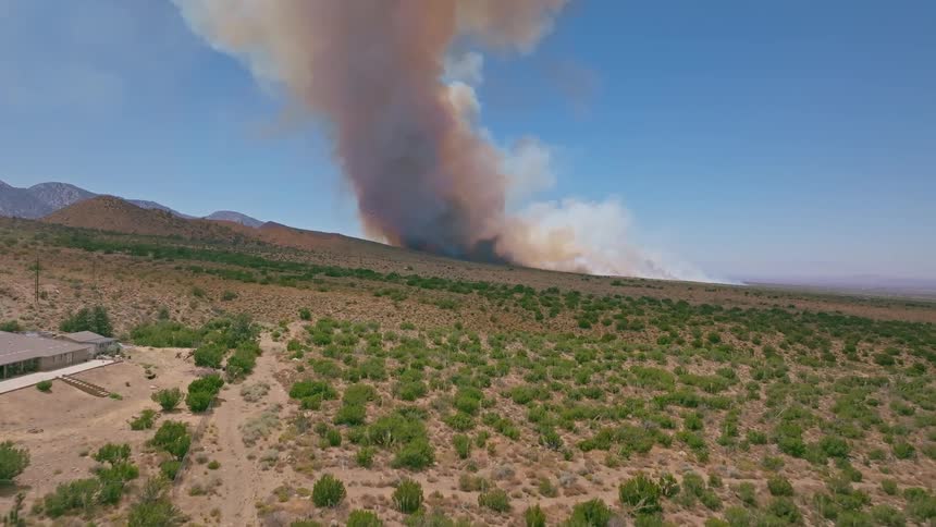 An aerial view shows a firefighting plane dropping red fire retardant on a massive wildfire burning through a dry, brushy landscape near mountains