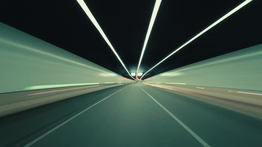 Futuristic POV time-lapse of a car speeding through a city at night and entering a brightly lit, modern tunnel