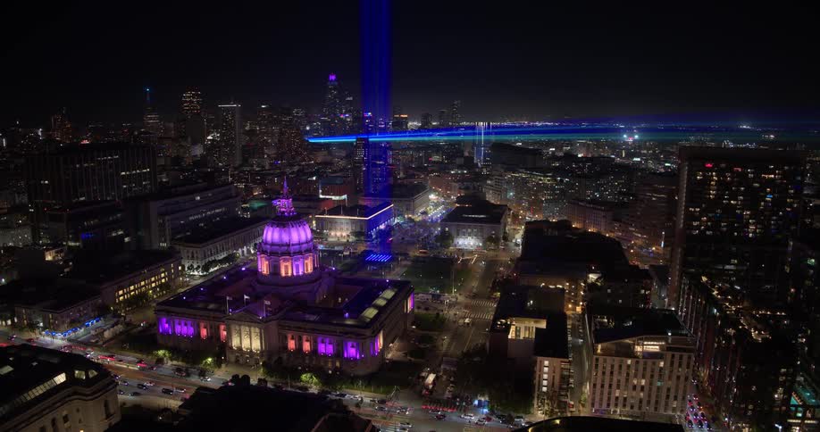 Stunning aerial time-lapse captures San Francisco's City Hall illuminated in purple at night, with traffic streaks and a vibrant blue light beam against the city skyline