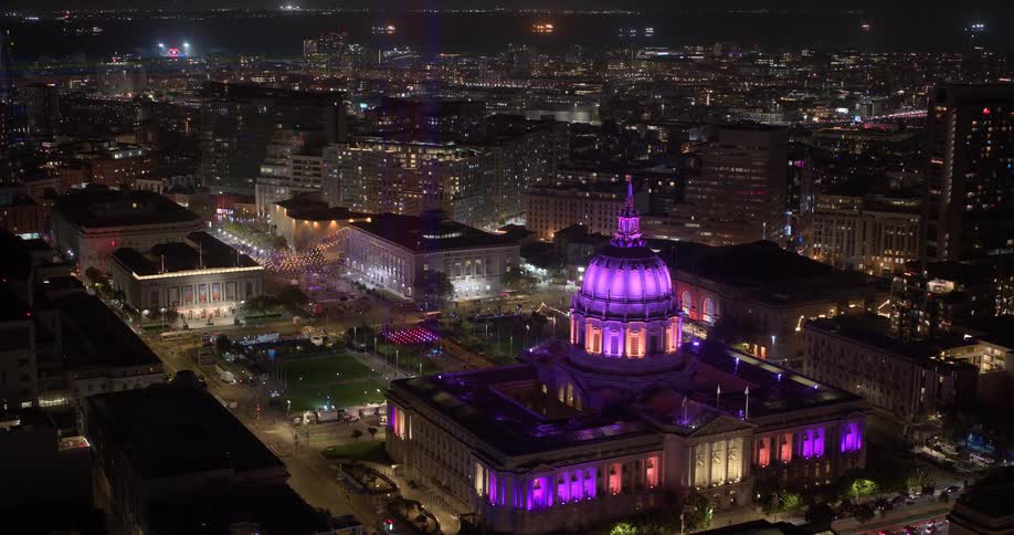 Cinematic aerial shot slowly pushing in on the San Francisco City Hall, brilliantly lit in purple and pink, amidst the glowing city at night