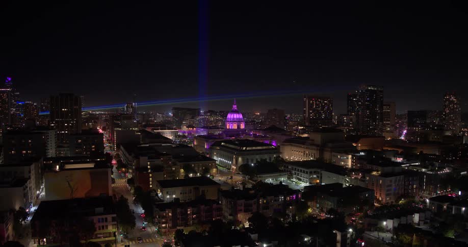 A breathtaking aerial view of San Francisco City Hall illuminated in vibrant purple at night, with a spectacular laser light show beaming across the urban skyline