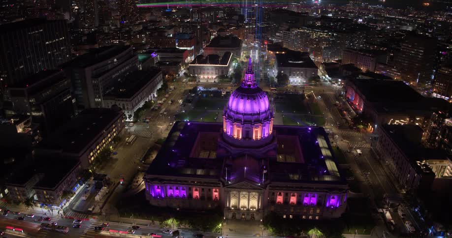 Spectacular aerial view pulls back from the iconic San Francisco City Hall, brightly illuminated in purple light against the dazzling downtown skyline at night