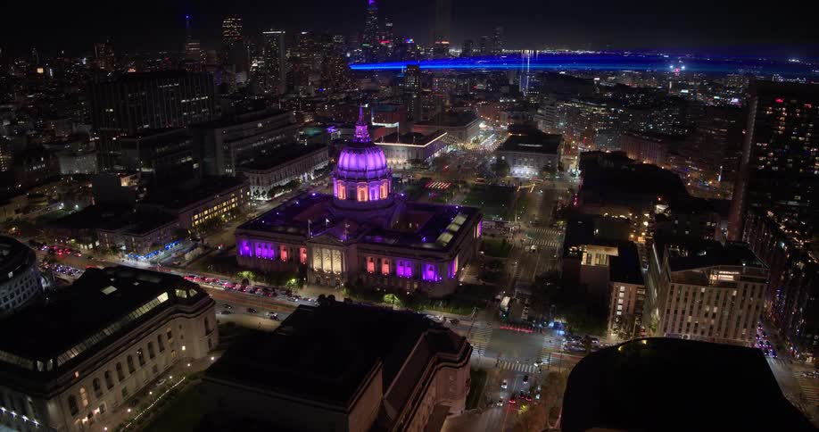 Spectacular aerial time-lapse view of San Francisco's illuminated City Hall at night, with a powerful light beam emerging from its dome over the bustling city