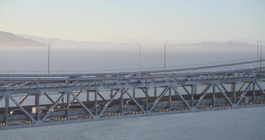 A wide shot shows traffic moving across both decks of the Oakland Bay Bridge in San Francisco during a hazy sunset