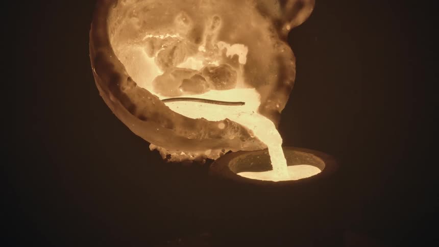A glowing, white-hot molten metal is carefully poured from a crucible into a mold in a dark foundry