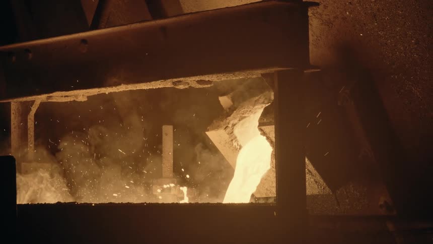 A stream of molten metal pours from industrial machinery in a dark foundry, creating smoke and sparks