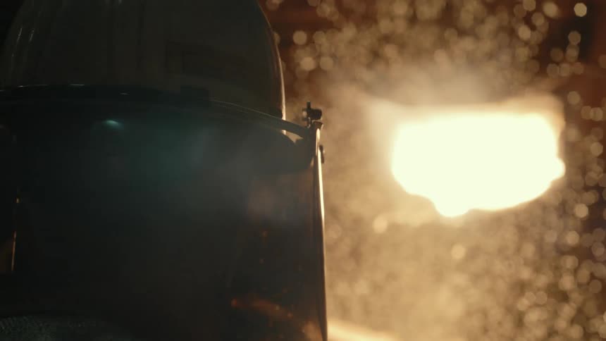 Cinematic close-up of a foundry worker's protective helmet with sparks and smoke billowing from a molten metal furnace in the background