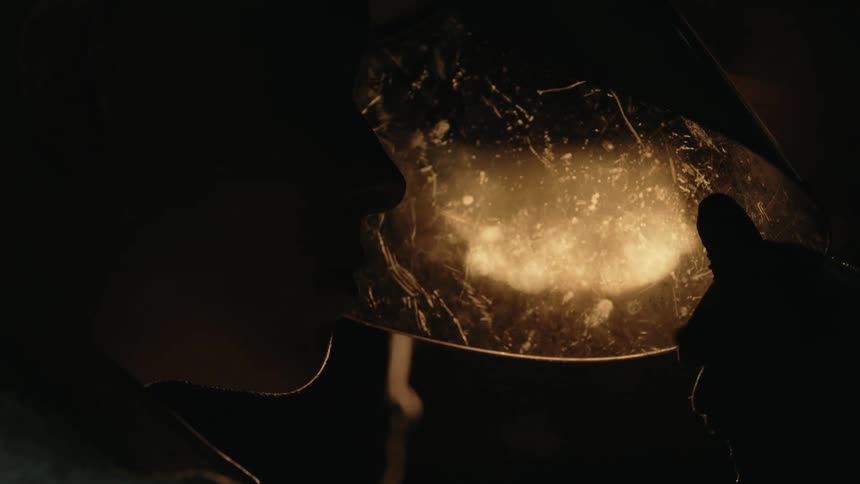 A craftsman in silhouette carefully inspects the sharp blade of a knife in a dark, moody workshop
