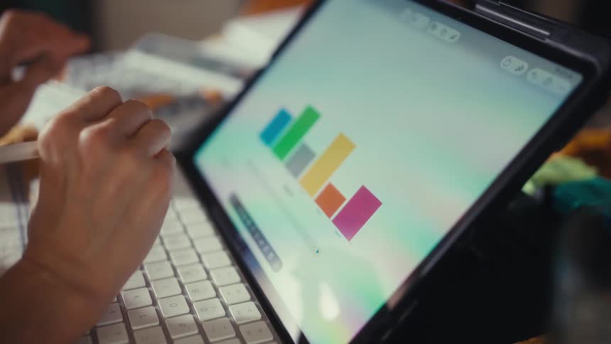 A person uses a stylus to analyze a colorful bar chart on a tablet while another person calculates figures in the background