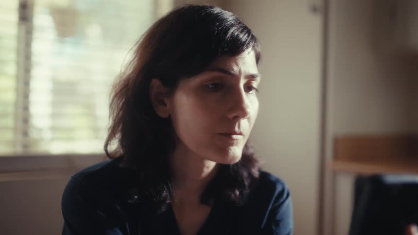 A serious woman with a pensive expression looks at her smartphone screen in a softly lit room