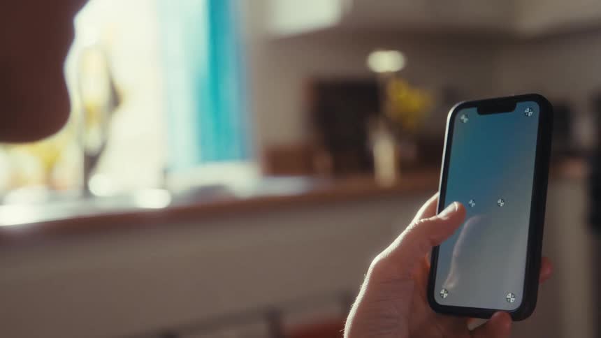 An over-the-shoulder view of a person holding and scrolling on a smartphone with a blank screen for compositing, inside a sunlit home