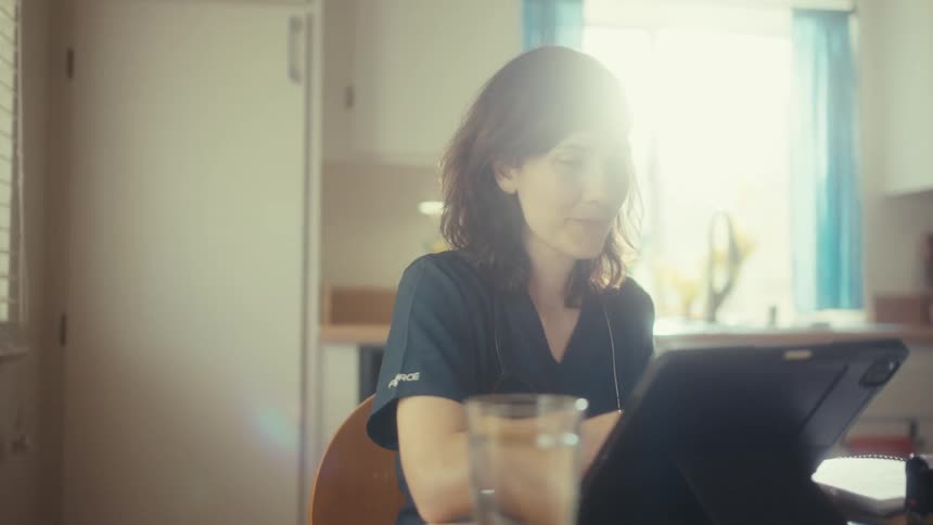 A young nurse in scrubs sits at her kitchen table in the morning sun, working diligently on a tablet