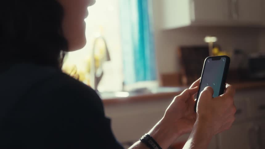 An over-the-shoulder shot captures a woman scrolling on her smartphone in a bright, sunlit kitchen