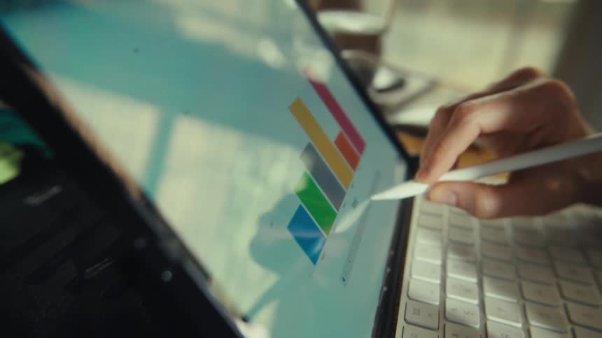 Close-up of a person's hand using a stylus to analyze a colorful bar chart on a tablet with a keyboard