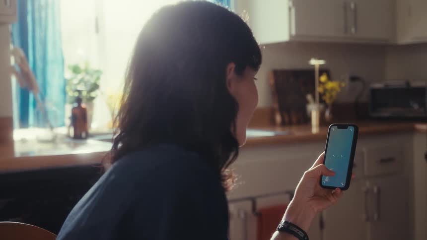 An over-the-shoulder view shows a woman holding a smartphone and interacting with the screen in a sunlit kitchen