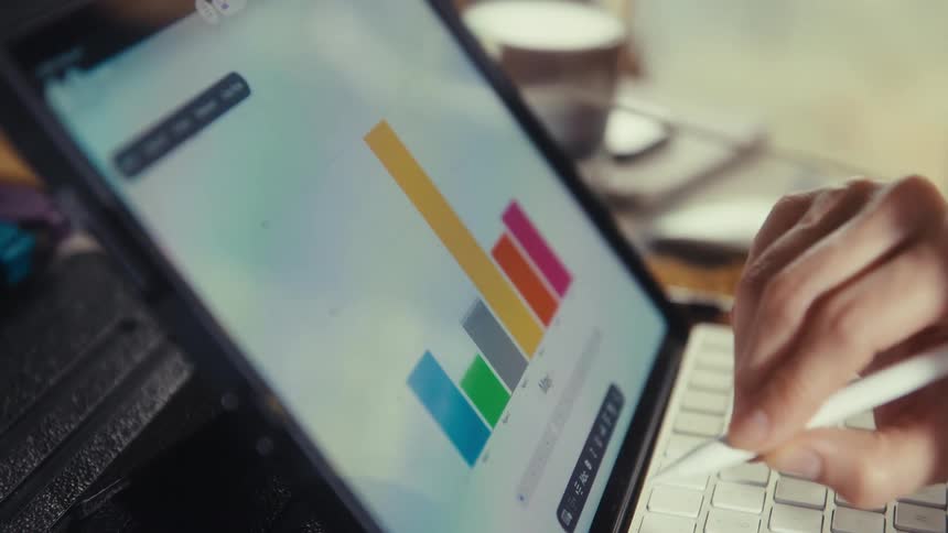 A person's hand uses a stylus to analyze a colorful bar chart on a tablet while working at a desk