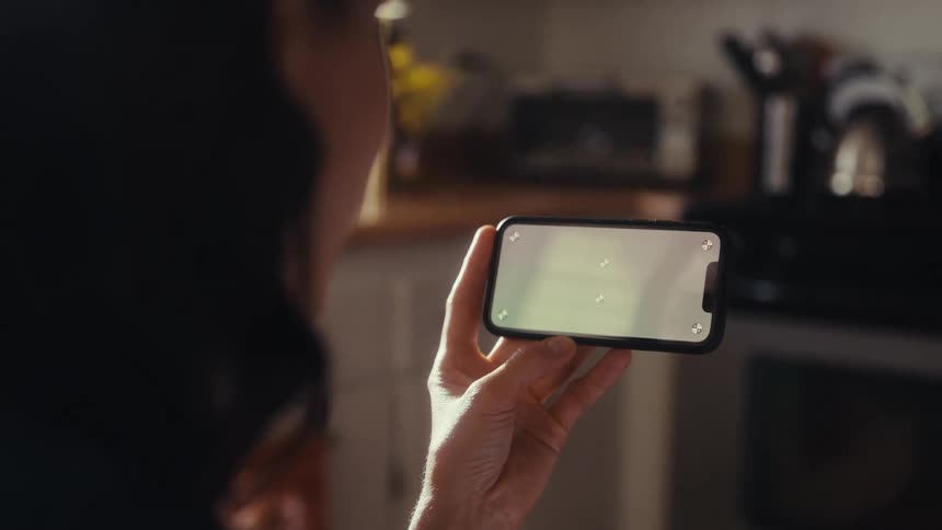 An over-the-shoulder shot of a woman holding a smartphone horizontally with tracking markers on the screen inside her kitchen
