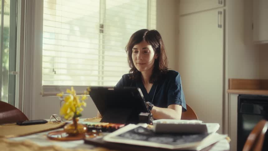 A woman in blue scrubs uses a stylus to draw on a tablet at her kitchen table