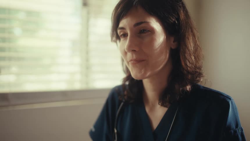 A caring female doctor in scrubs smiles warmly while listening to a patient in her naturally lit office