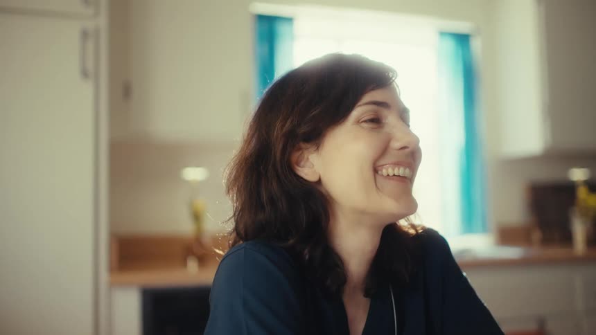 Slow motion shot of a happy female nurse in scrubs laughing during a video call on her tablet in a sun-drenched kitchen