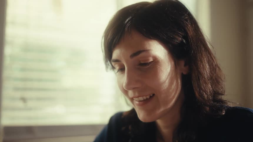 A female doctor in scrubs smiles thoughtfully while looking out a window in her sunlit office