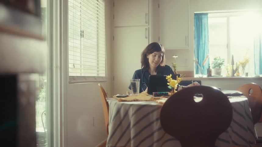 A female doctor in scrubs smiles while working on her tablet at her dining room table in a sunlit kitchen