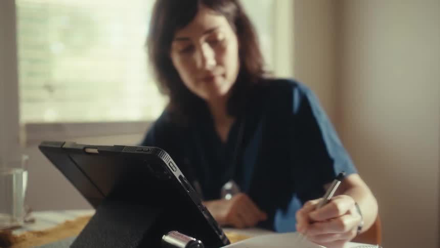 A rack focus shot reveals a dedicated female doctor in scrubs studying with a tablet, with medical scans and a calculator on the table in the foreground