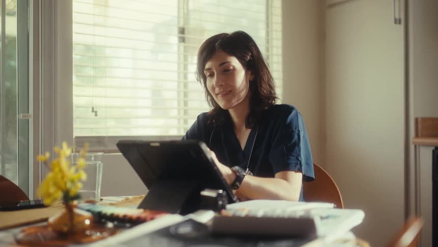 A thoughtful female doctor in scrubs finds a moment of creative peace, drawing on her digital tablet at her sunlit kitchen table